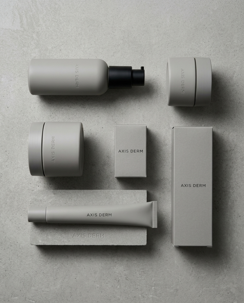 Matte warm grey Axis Derm skincare set: pump bottle, two jars, tube and two boxes on a concrete surface.